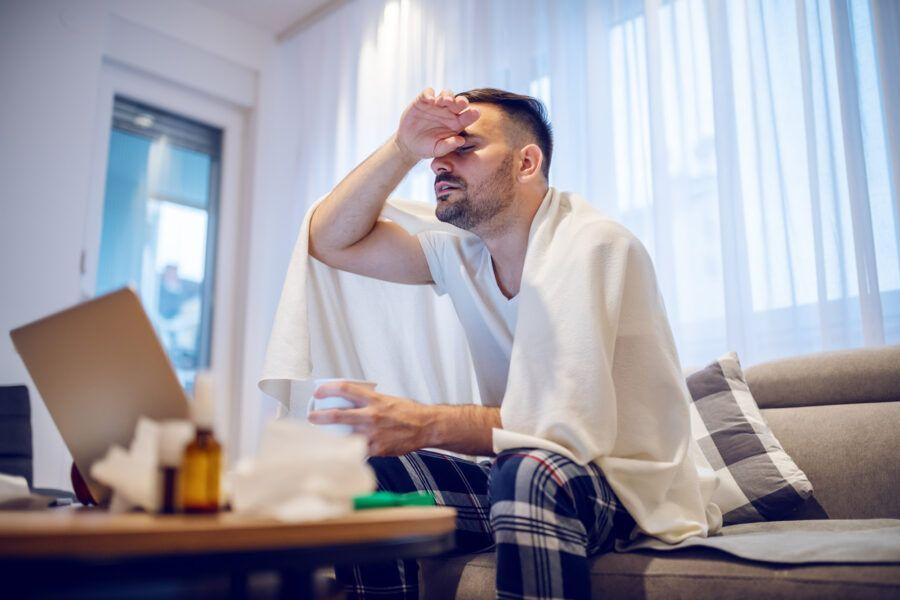 Hardworking employee in pajamas and covered with blanket having fever. Man holding mug with tea and sitting on sofa in living room.