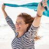 Smiling mature woman holding blue scarf over head while walking at beach. Happy active and healthy woman enjoying summer vacation at sea. Middle aged lady running at seashore with blue fabric.